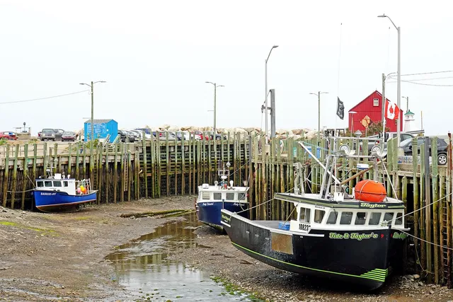 Fishing boats resting on the muddy harbour floor at low tide beside tall timber wharf pilings at Hall's Harbour, Nova Scotia, with a red waterfront building and a Canadian flag behind