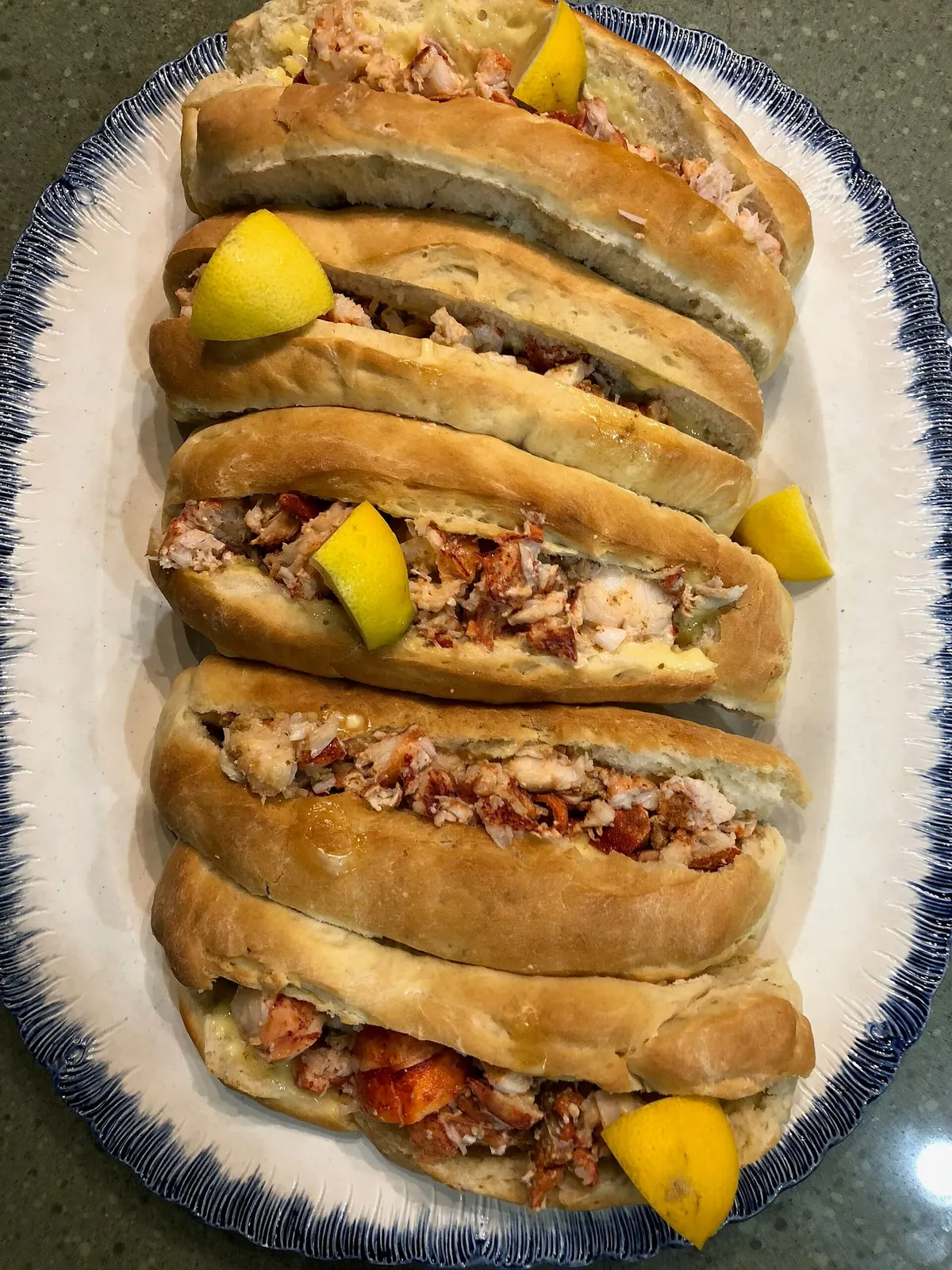 Five homemade lobster rolls on an oval platter with a blue rim, each bun packed with chunks of pink-and-white lobster meat and garnished with a lemon wedge