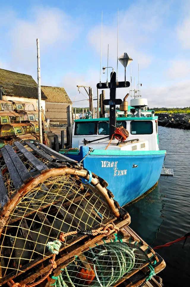Wooden lobster traps with coloured tags stacked on the wharf at West Berlin, Queens County, beside the blue and white lobster boat Wade-N-Lynn, with weathered fish sheds behind